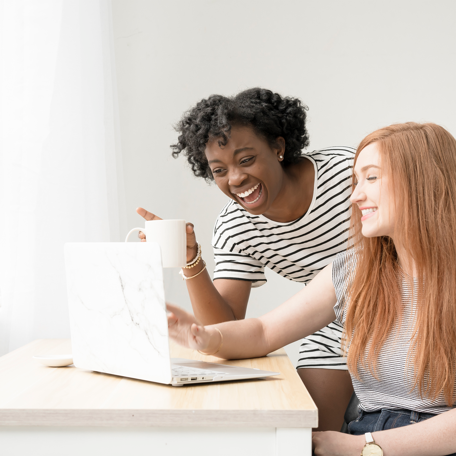 Black person in black and white striped short sleeve top laughing with white mug in hand next to a white red-haired person with gray & white striped cap sleeved shirt sitting at a white & gold marble laptop. learning product safety compliance