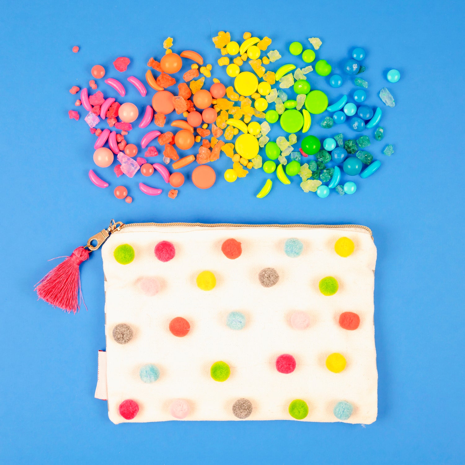 White fabric snack bag with colorful pompoms on the sides, a pink tassle on the gold zipper pull and a rainbow of candies spread above the bag on a blue background.